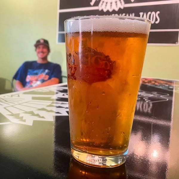A pint of beer with a foamy head on a table, a smiling person in the background.