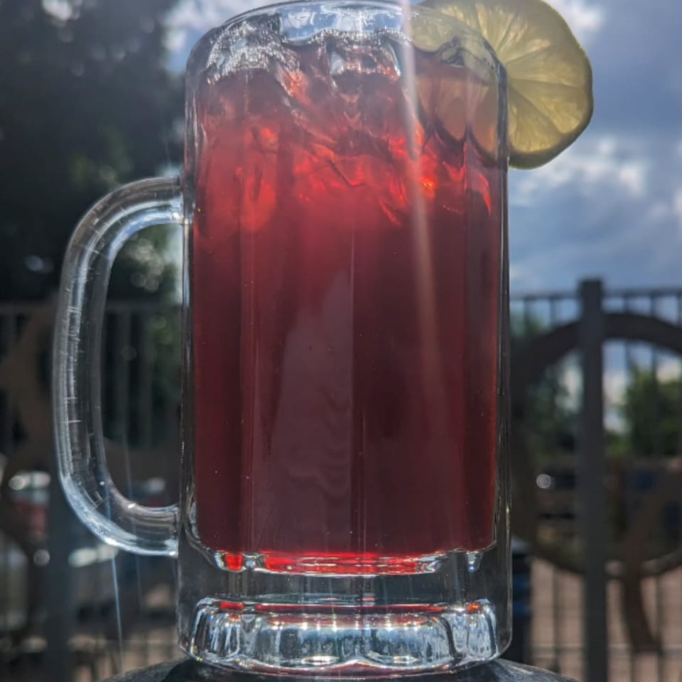 A large glass mug of DGT Tacos' red agua fresca with ice and a lemon slice, outdoors.