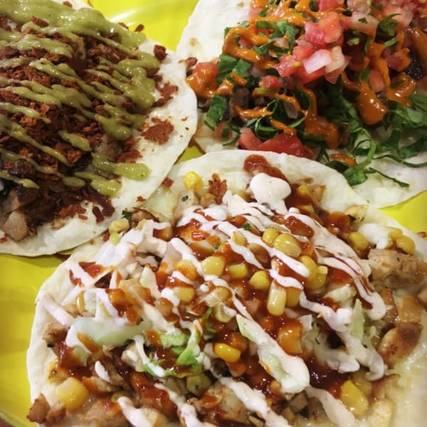 Three DGT Tacos on a yellow plate, topped with various meats, sauces, and fresh ingredients.