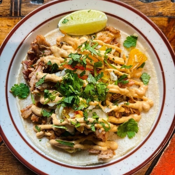 A close-up of a taco with meat, sauce, cilantro, and a lime wedge on a white plate.